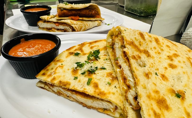 Two plates of chicken quesadillas with melted cheese, garnished with parsley, and served with red dipping sauce.