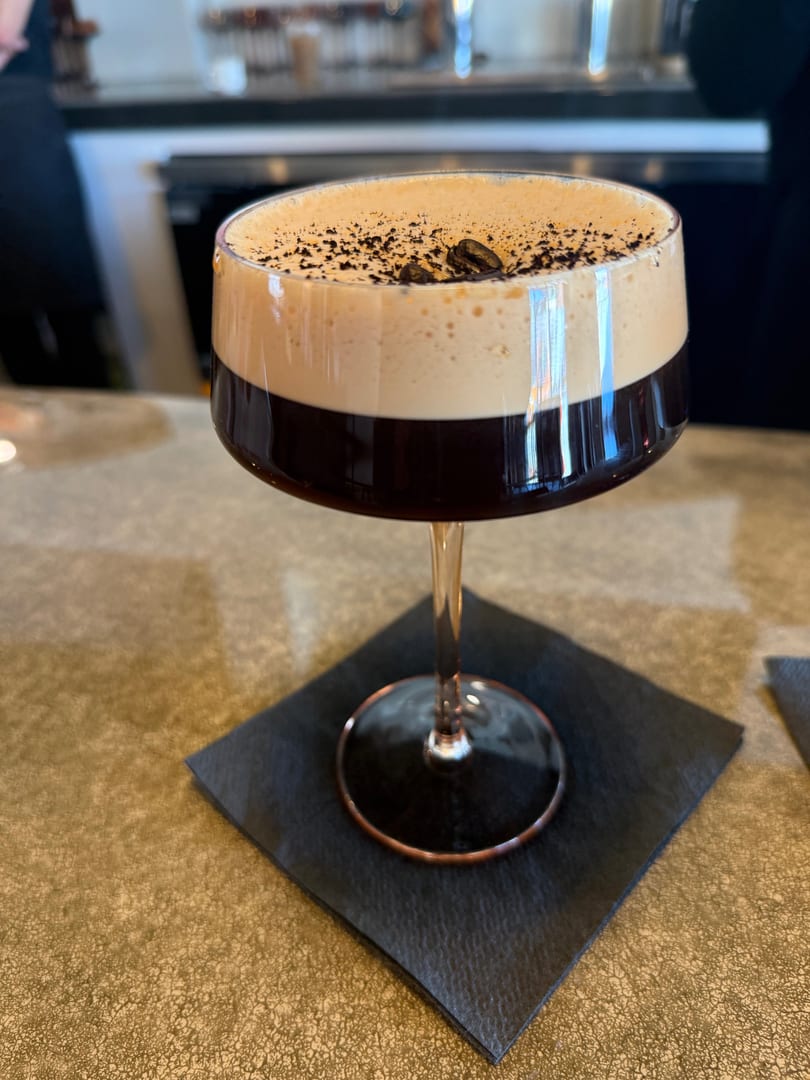 Espresso martini in a coupe glass with foamy top, coffee beans, and cocoa powder on a black napkin.